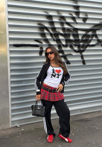 Mujer con chaqueta negra, camiseta blanca con "I love NY", falda roja de cuadros sobre pantalones negros, zapatillas rojas, gafas de sol, sosteniendo bolso negro, posando junto a pared con grafiti.
