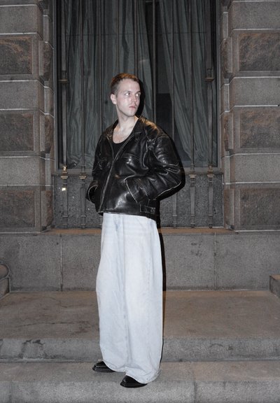 Chaqueta bomber de cuero negra, vaqueros de denim claros y sueltos, zapatos negros. El fondo presenta escalones de piedra y cortinas colgadas. Iluminación de atardecer.