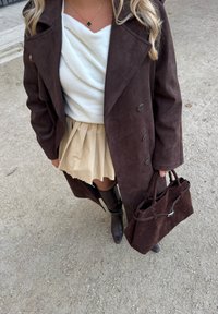 Brun semsket frakk med store knapper, hvit drapert topp, plissert beige skjørt og brune knelange støvler, holder en matchende brun toteveske.