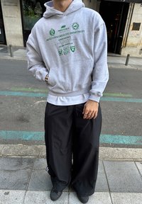 Grå hettegenser med grønne grafiske detaljer, en kengurulomme og ribbestrikkede mansjetter, kombinert med løse svarte bukser med vide ben, brukt med svarte joggesko.