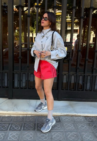 Mujer con gafas de sol, chaqueta gris de gran tamaño, pantalones cortos rojos, zapatillas grises, sosteniendo una bebida, de pie frente a una puerta de metal negra en la acera.