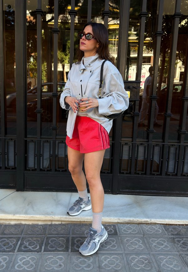 Femme avec des lunettes de soleil, veste grise oversize, short rouge, baskets grises, tenant un verre, debout devant une grille en métal noire sur le trottoir.