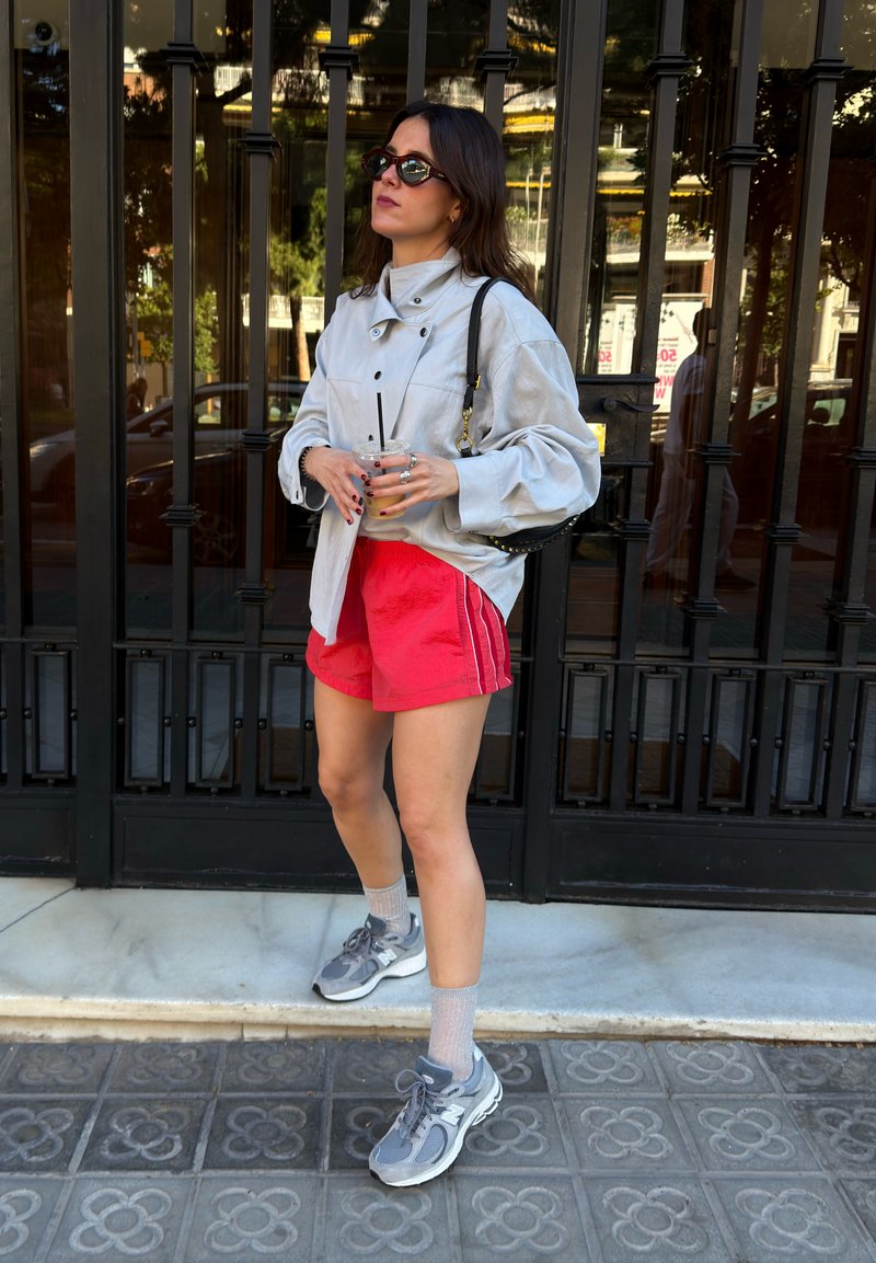 Femme avec des lunettes de soleil, veste grise oversize, short rouge, baskets grises, tenant un verre, debout devant une grille en métal noire sur le trottoir.