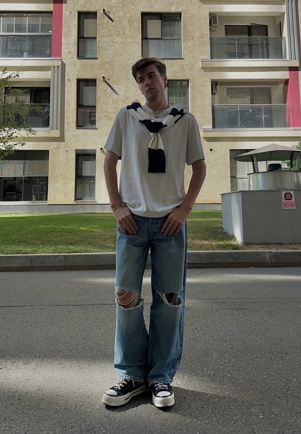 Camisa branca de manga curta com um cachecol escuro à volta do pescoço, jeans azul claro desgastados e ténis pretos com solas brancas. Fundo urbano.