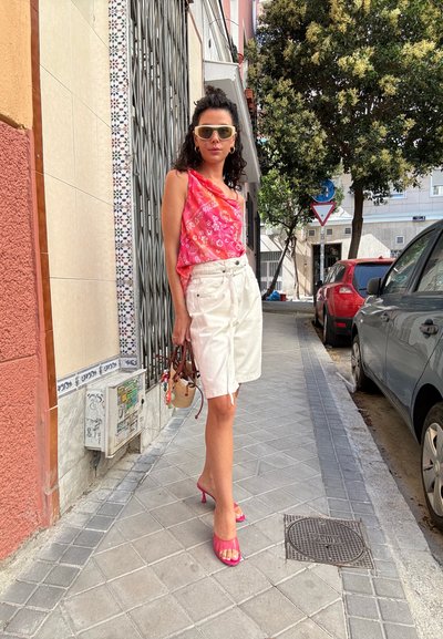 Top sin mangas de color rosa con estampado, pantalones cortos blancos, tacones abiertos rosa. Lleva una bolsa pequeña con charms coloridos. Calle pavimentada y edificios al fondo.