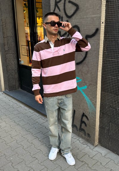 Polo de manga larga a rayas marrón y rosa con cuello; vaqueros azul claro; zapatillas blancas; de pie frente a una pared texturizada.