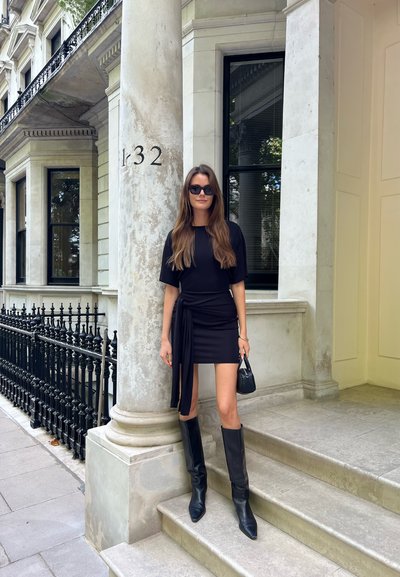 Mujer con vestido negro y botas, llevando un bolso, de pie en escalones de piedra junto a un pilar marcado con "132" fuera de un edificio clásico de piedra.
