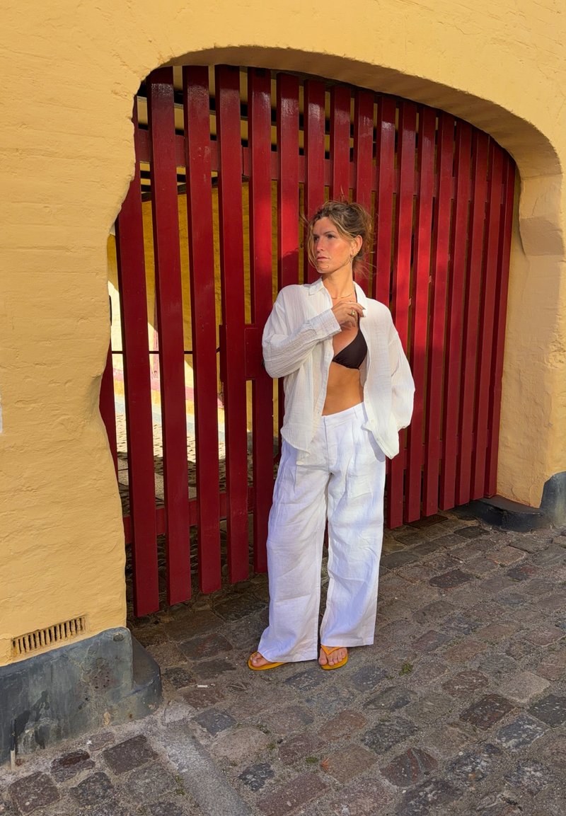 Femme en pantalon blanc ample et chemise sur un haut noir, debout dans une rue pavée près d'un mur jaune et d'une grande porte en bois rouge.