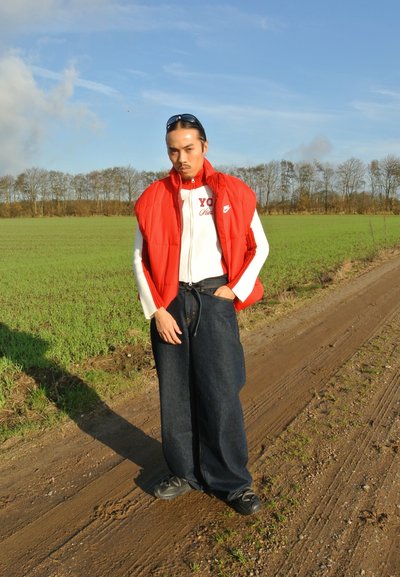 Chaleco acolchado rojo sobre una camiseta blanca con cremallera que tiene el texto "YO", combinado con pantalones vaqueros oscuros holgados y zapatillas negras, de pie en un camino de tierra.