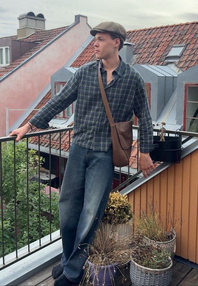 Joven con una camisa a cuadros verde y azul marino, jeans de mezclilla y un bolso bandolera marrón, de pie en un balcón con plantas en macetas.