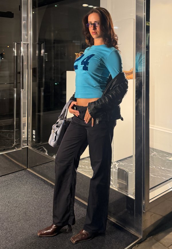Mujer con gafas que lleva una camiseta corta azul con el número 24, pantalones negros y sostiene una chaqueta negra y un bolso gris junto a puertas de cristal.