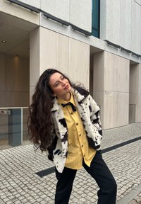 Mākslīgās kažokādas mētelis ar melnbaltu govs rakstu, dzeltena pogām aizsegta kreklē, melns tauriņs un melnas bikses, ar pūkainu tekstūru un vaļīgu piegriezumu.