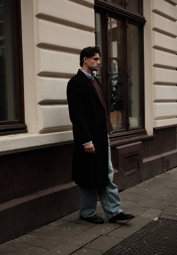 Uomo con lungo cappotto nero, maglione marrone e jeans blu in piedi su un marciapiede cittadino vicino a un edificio bianco con ampie finestre.