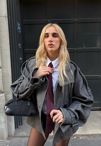 Blazer grigio oversize a motivo pied de poule con maniche larghe, camicia bianca e cravatta bordeaux; tiene una piccola borsa nera.
