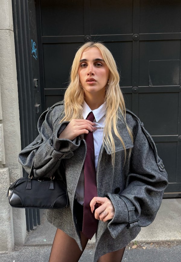 Blazer grigio oversize a motivo pied de poule con maniche larghe, camicia bianca e cravatta bordeaux; tiene una piccola borsa nera.