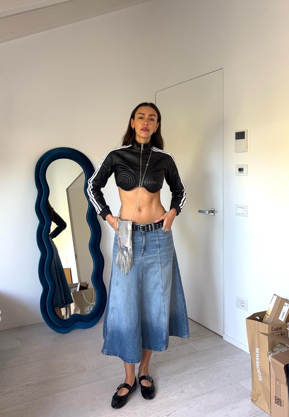 Czarna krótka kurtka zapinana na zamek z spiralnym wzorem, niebieska midi spódnica z dżinsu, srebrna torba na pasek i czarne buty. W tle lustro refleksyjne.