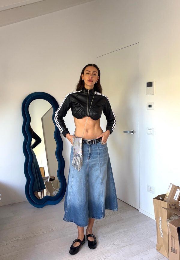 Giubbotto nero corto con zip e design a spirale, gonna midi in denim blu, borsa a cintura argentata e scarpe nere. Specchio riflettente sullo sfondo.