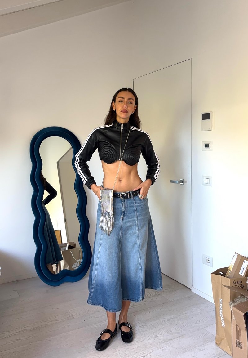 Schwarze, verkürzte Zip-Up-Jacke mit Spiraldesign, bleu denim Midi-Rock, silberne Gürteltasche und schwarze Schuhe. Reflexionsspiegel im Hintergrund.