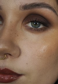 Nærbilde av et ansikt med lys brun hud, fremtredende fregner, gull septumring, brun øyenskygge og glinsende mørkerøde lepper.