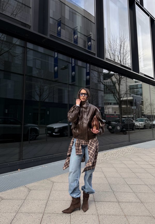 Žena nosi nadrozmernú hnedú koženú bundu, károvanú košeľu zviazanú v páse, svetlomodré džínsy, hnedé topánky, slnečné okuliare, drží zladenú tašku na mestskom chodníku.