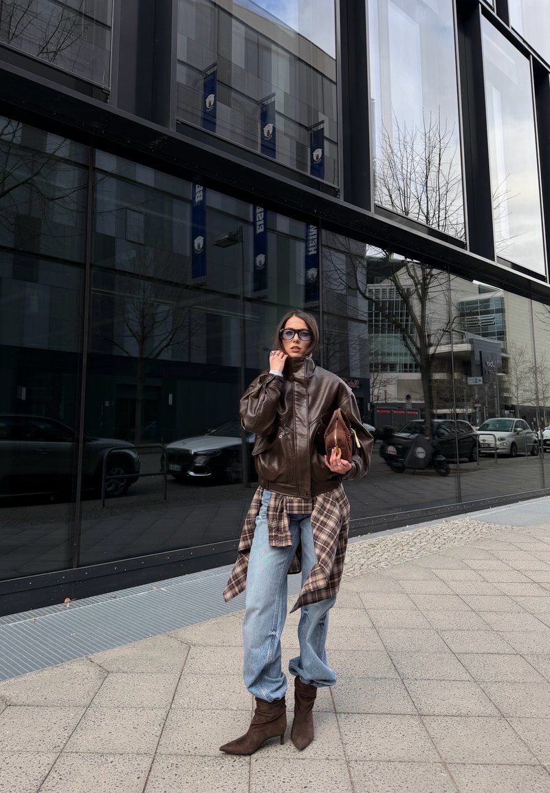 Femme portant une veste en cuir marron surdimensionnée, chemise à carreaux nouée à la taille, jean bleu clair, bottes marron, lunettes de soleil, tenant un sac assorti sur le trottoir en ville.