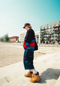 Černá a červená bunda s modrými pruhy, modré džínové šortky s červenou výšivkou a béžové boty s tkaničkami. Venkovní městské prostředí.