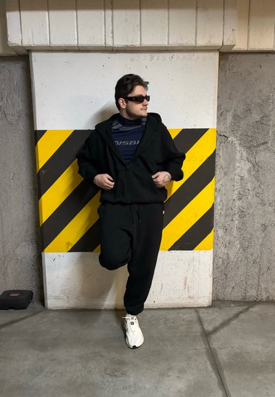 Sudadera negra con cremallera combinada con pantalones de chándal negros ajustados. Los accesorios incluyen gafas de sol oscuras y zapatillas deportivas blancas. Pared distintiva de rayas amarillas y negras detrás.