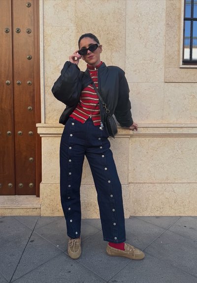 Chaqueta acolchada negra, camiseta de rayas rojas y blancas, pantalones de mezclilla azules con detalles de botones plateados y zapatos beige con calcetines rojos. De pie sobre el pavimento.