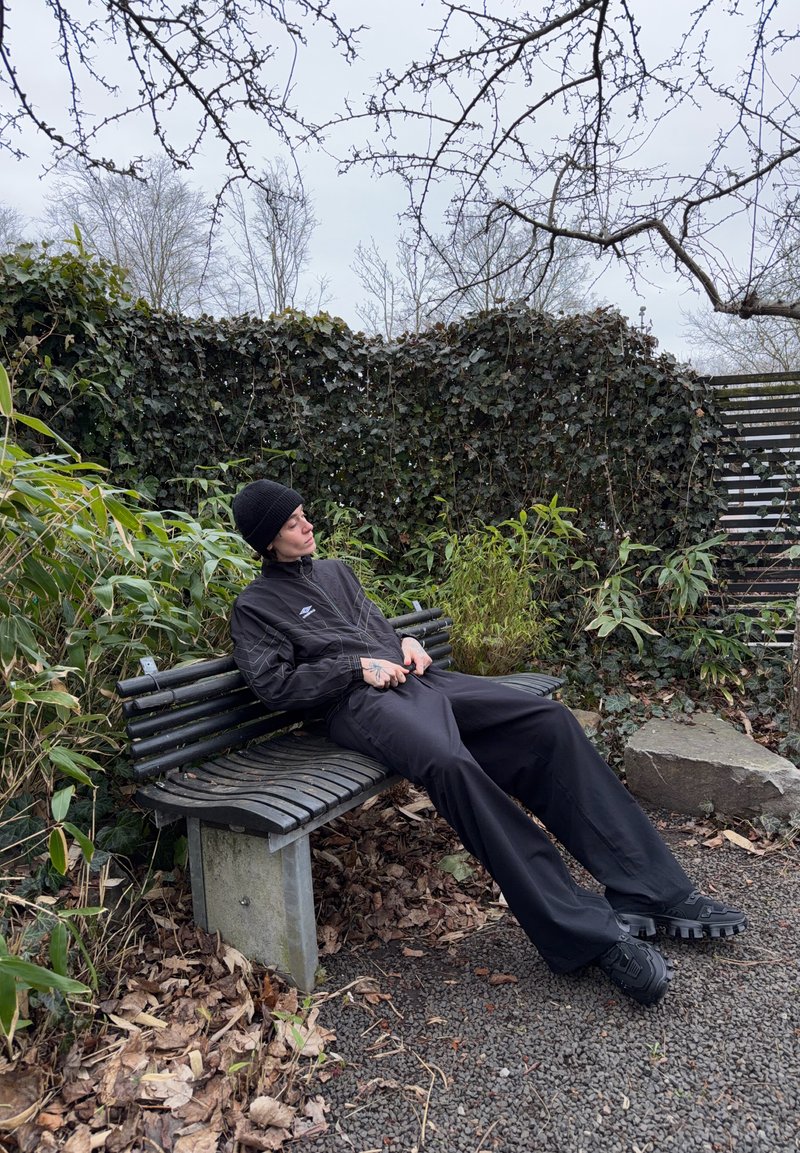 Person in black clothing and hat sitting and leaning back on a curved wooden park bench surrounded by plants and fallen leaves.