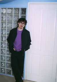 Mladi odrasel moški stoji ob steni s steklenimi bloki. Oblečen je v črno jopa z zadrgo, vijolično majico in temne hlače. Za njim je bela vrata.