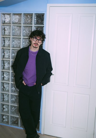 Joven adulto de pie contra una pared de bloques de vidrio. Lleva una chaqueta negra con cremallera, una camiseta morada y pantalones oscuros. Puerta blanca detrás.