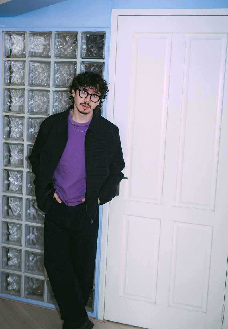 Young adult man stands against a wall with glass blocks. He wears a black zip-up jacket, purple shirt, and dark pants. White door behind.
