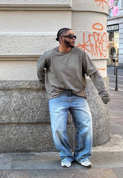 Sudadera de manga larga color oliva, con un corte relajado, combinada con jeans de mezclilla azul holgados y zapatillas blancas, frente a una pared texturizada de color beige.