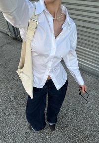 Biała koszula na guziki, luźne ciemne dżinsy, kremowa torba na ramię z suwakiem oraz czarne buty. Tło ma metaliczną teksturę.