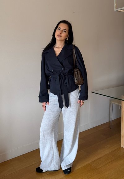 Chaqueta azul marino de doble botonadura con cintura ajustable, combinada con pantalones de chándal de pierna ancha en gris claro. También incluye un bolso marrón y pantuflas negras.