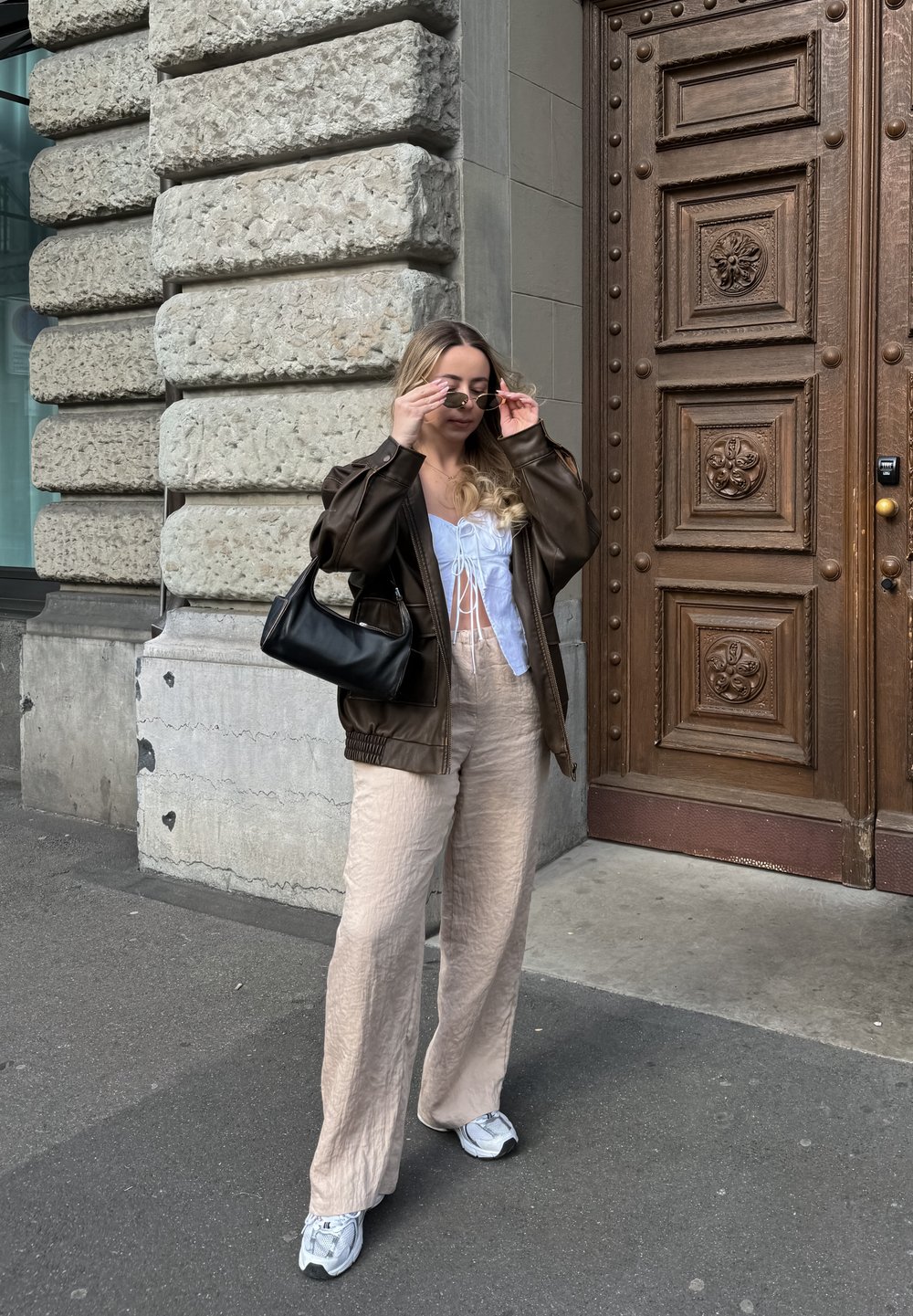 Καφέ δερμάτινο υπερμεγέθες τζάκετ, λευκή μπλούζα με κορδόνια, μπεζ υφασμένα παντελόνια σε φαρδιά γραμμή, μαύρη τσάντα, γκρι αθλητικά παπούτσια, στέκεται μπροστά σε τοίχο από πέτρα.