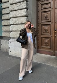 Καφέ δερμάτινο υπερμεγέθες τζάκετ, λευκή μπλούζα με κορδόνια, μπεζ υφασμένα παντελόνια σε φαρδιά γραμμή, μαύρη τσάντα, γκρι αθλητικά παπούτσια, στέκεται μπροστά σε τοίχο από πέτρα.