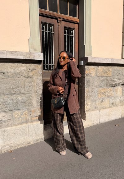 Mujer con chaqueta marrón, pantalones a cuadros, gafas de sol y zapatos beige de pie frente a una puerta de madera con fondo de muro de piedra.
