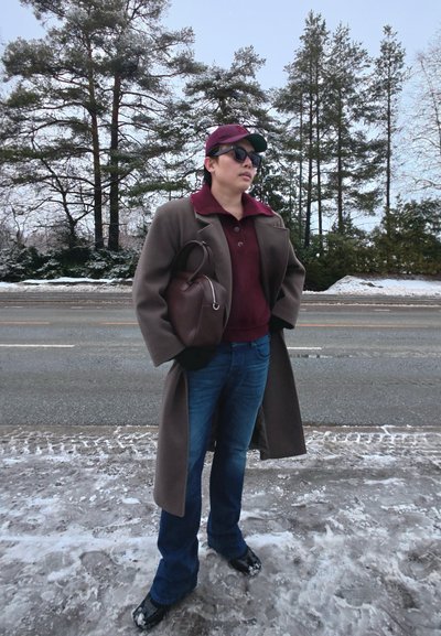 Persona con gafas de sol, gorra marrón, abrigo y vaqueros, de pie en el borde de una carretera nevada con árboles al fondo, sosteniendo un bolso marrón.