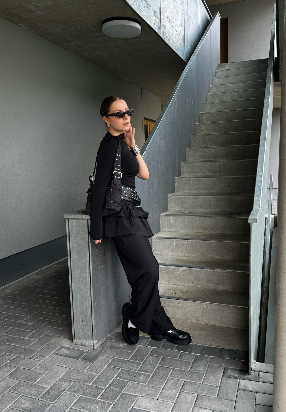 Μαύρο στιβαρό σύνολο με μακριά μανίκια, λεπτομέρειες και υφασμάτινη φούστα. Στέκεται δίπλα σε μια σκάλες από τσιμέντο, φορώντας μαύρα γυαλιά ηλίου και παπούτσια.