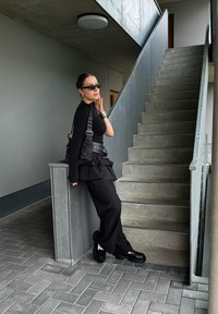 Μαύρο στιβαρό σύνολο με μακριά μανίκια, λεπτομέρειες και υφασμάτινη φούστα. Στέκεται δίπλα σε μια σκάλες από τσιμέντο, φορώντας μαύρα γυαλιά ηλίου και παπούτσια.