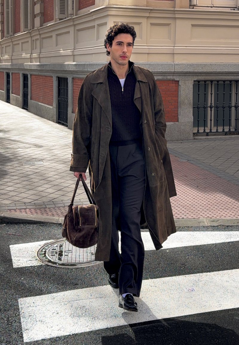 Homme marchant sur un passage piéton en ville, portant un long manteau sombre, un pantalon noir et portant un sac en cuir marron.