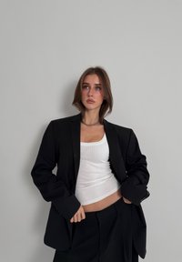 Blazer noir ajusté porté sur un débardeur blanc côtelé, avec une coupe ample et un pantalon noir à jambes larges, sur un fond neutre.
