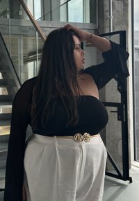 Femme aux longs cheveux foncés portant un haut noir à manches longues et épaules dénudées, et un pantalon blanc taille haute avec une boucle de ceinture dorée en forme de fleur, à l'intérieur.