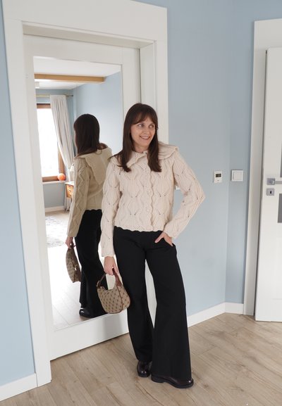 Mujer con suéter beige de punto y pantalones negros sosteniendo un bolso, de pie junto a un espejo de cuerpo entero en una habitación azul claro.