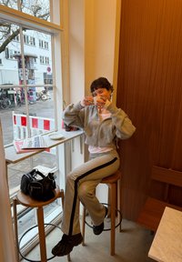 Personne en survêtement gris assise sur un tabouret en bois près d'une fenêtre, tenant une tasse blanche près du visage, avec des sacs sur un tabouret à proximité.