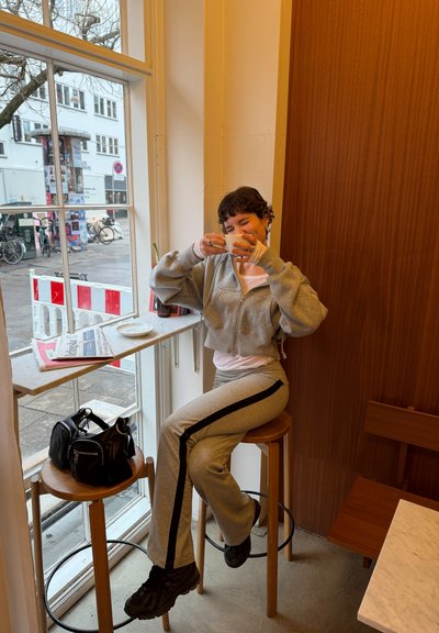 Persona con chándal gris sentada en un taburete de madera junto a una ventana, sosteniendo una taza blanca cerca de su rostro, con bolsas en un taburete cercano.