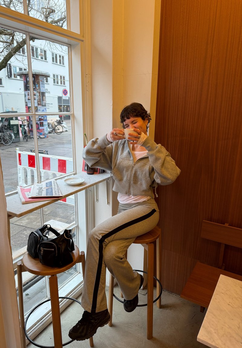 Person in grauem Trainingsanzug sitzt auf einem Holzstuhl am Fenster, hält eine weiße Tasse nahe am Gesicht, mit Taschen auf einem nahegelegenen Hocker.