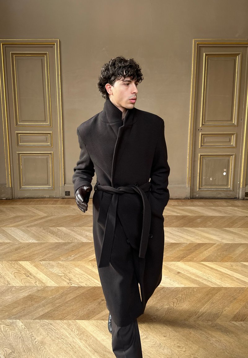 Man with curly hair wearing long black coat and glove, walking on wooden floor between two ornate beige doors.