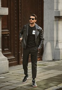 Fekete bőr motoros kabát fekete póló fehér logóval, testre szabott fekete nadrággal és fekete sportcipővel, fehér részletekkel párosítva.